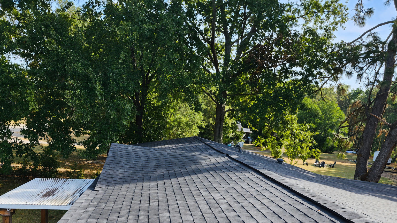 Charcoal architectural shingles across a roof we finished, photographed standing on it, oaks and yard beyond